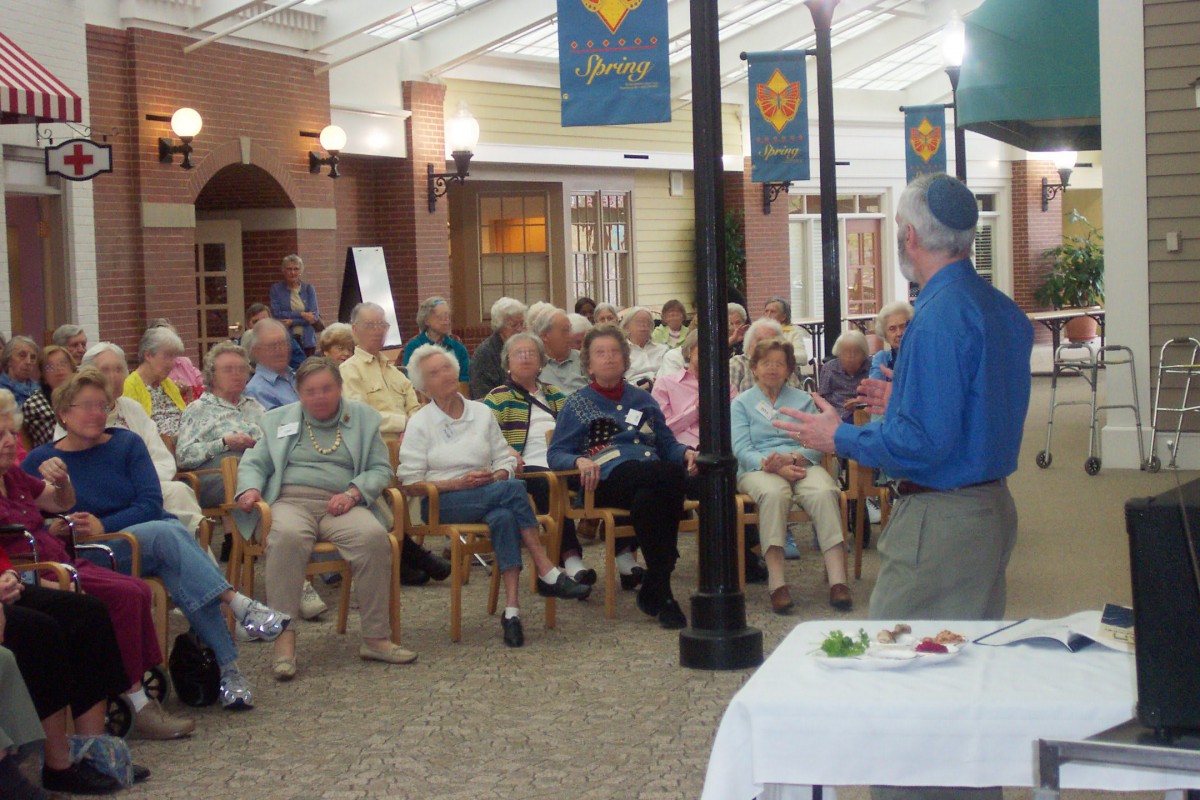 A speaker addresses a group of elderly people seated in an indoor community center setting.