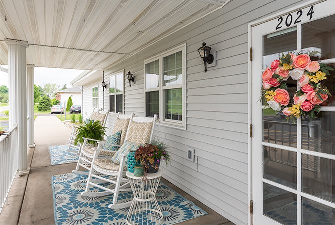 A cozy porch with white rocking chairs, colorful plants, and a decorative rug.