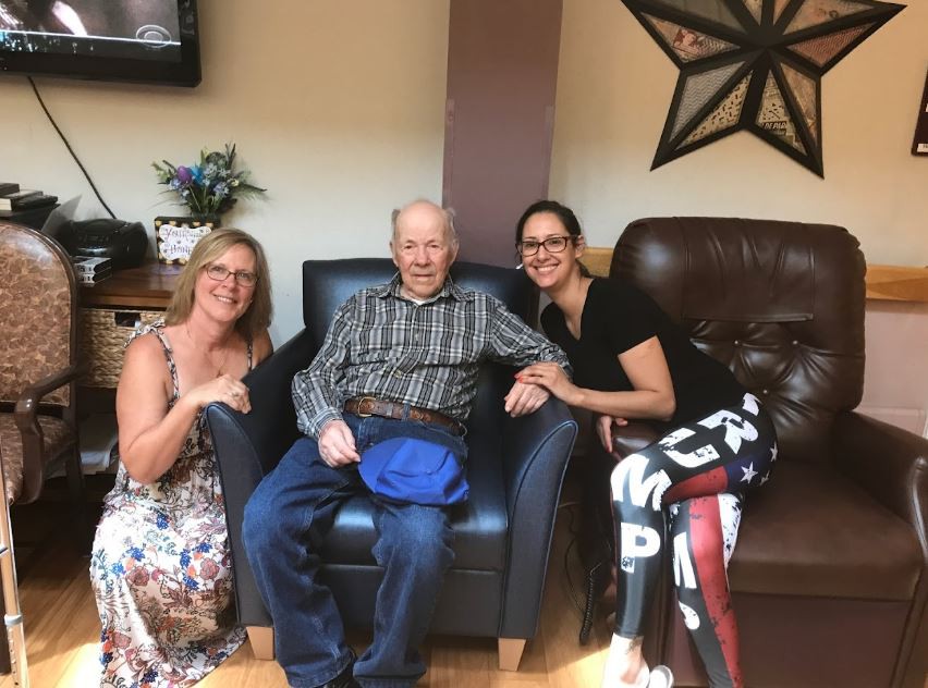 A group of three people posing together indoors, with an elderly person seated in the middle.