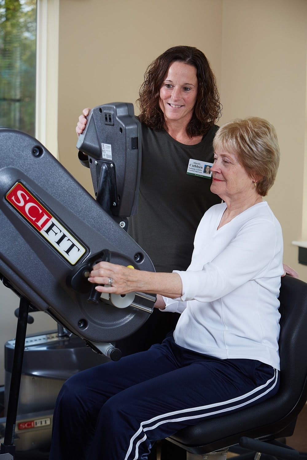 A woman assists another woman using a SCIFIT exercise machine for rehabilitation or fitness training.