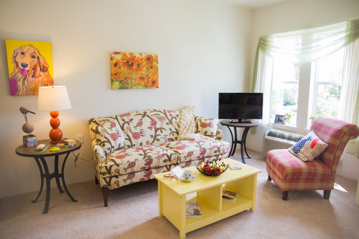 A cozy living room with a floral-patterned sofa, a yellow coffee table, and a television near the window.