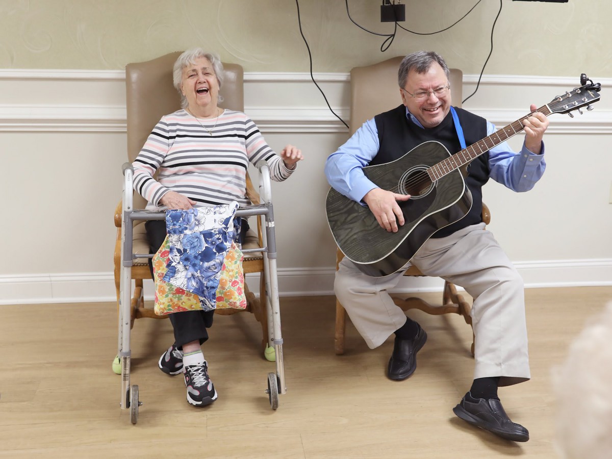 An elderly woman in a wheelchair enjoys listening to a man playing the guitar, both smiling and sharing a joyful moment.