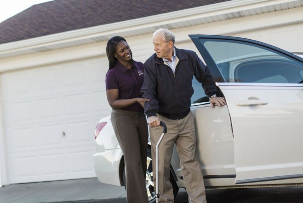 A caregiver assists an elderly man as he exits a car.