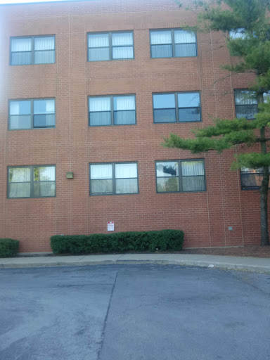 A brick building featuring multiple windows, surrounded by greenery and pavement.