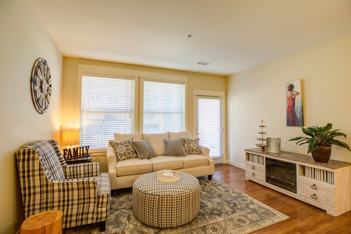 A cozy living room with a sofa, armchair, and ottoman arranged around a patterned carpet. Natural light filters through the large windows, creating a warm and inviting atmosphere.