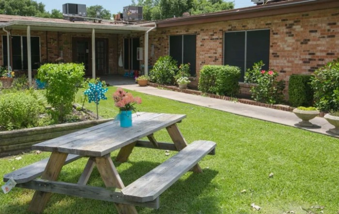 A picnic table is set on a lawn in front of a brick building, surrounded by lush greenery and flowers.