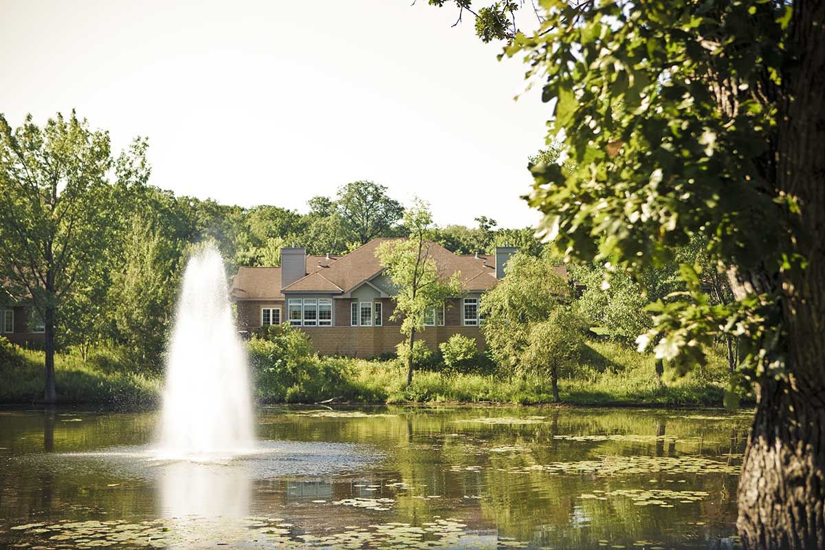 A serene landscape featuring a house by a pond with a fountain and surrounded by lush trees.