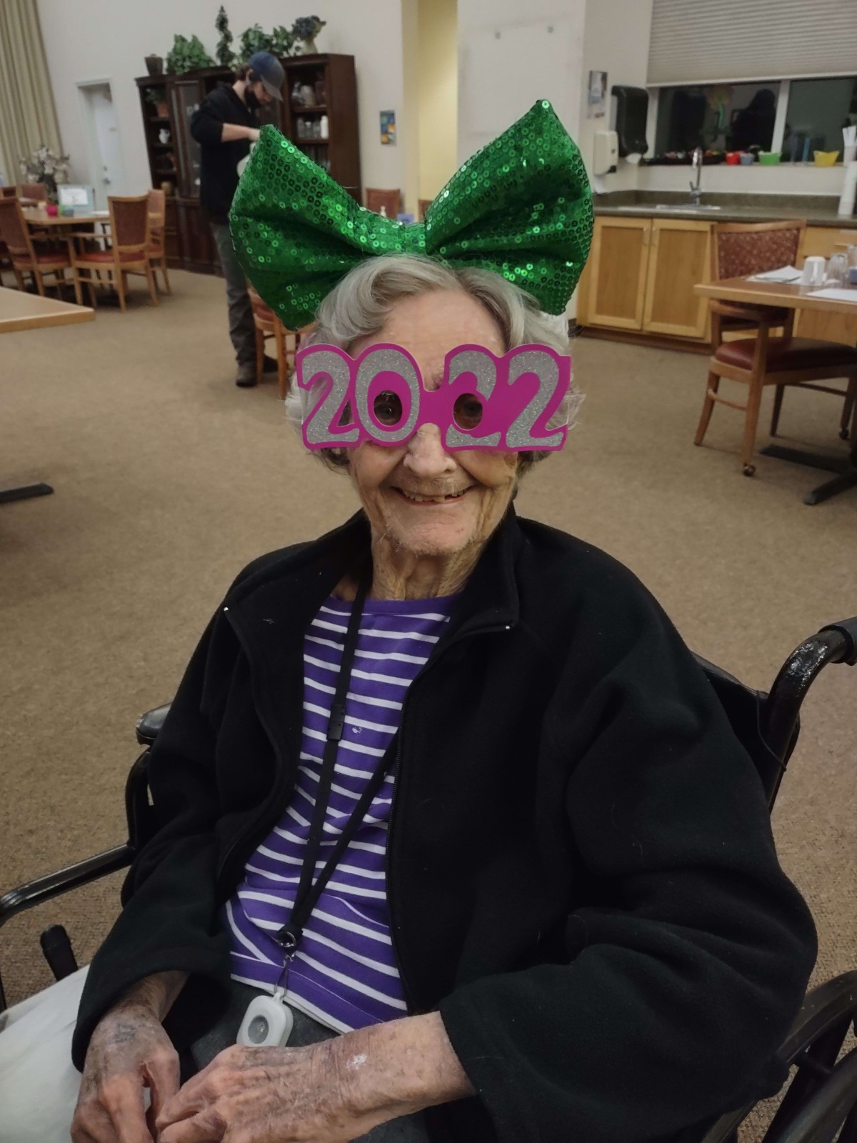 An elderly person in a wheelchair wearing festive 2022 glasses and a large green bow.