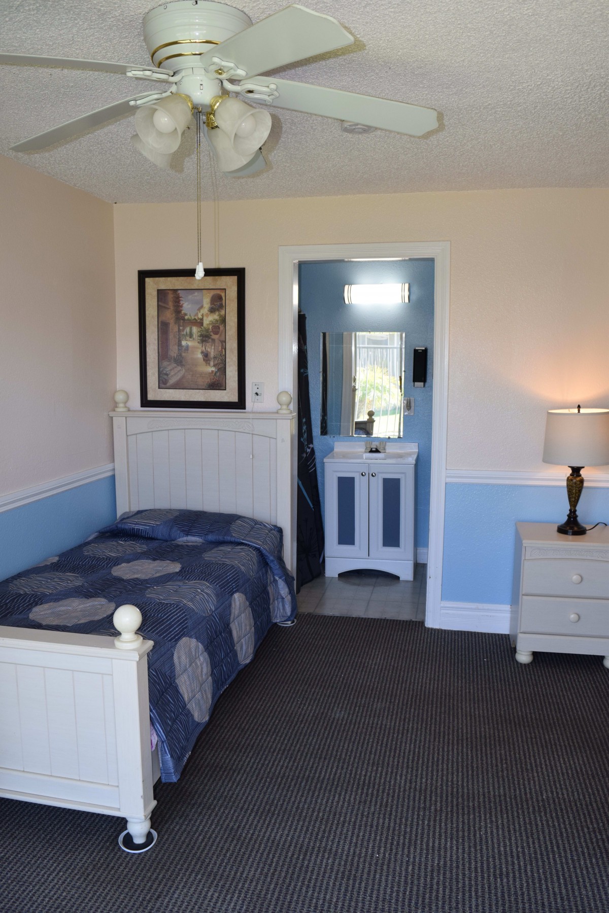 A cozy bedroom with a single bed, featuring blue and white decor, a nightstand with a lamp, and an adjoining bathroom.