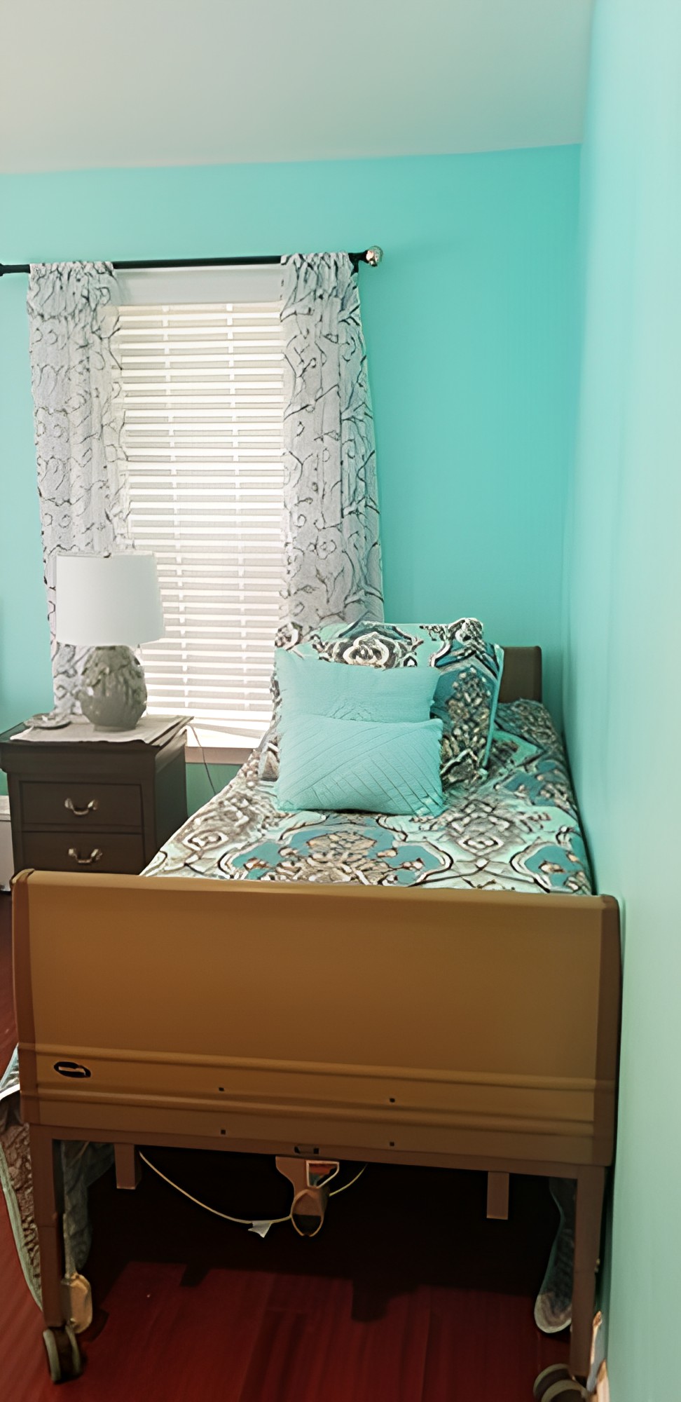 A cozy bedroom with turquoise walls and a single bed adorned with decorative pillows, next to a nightstand and lamp.