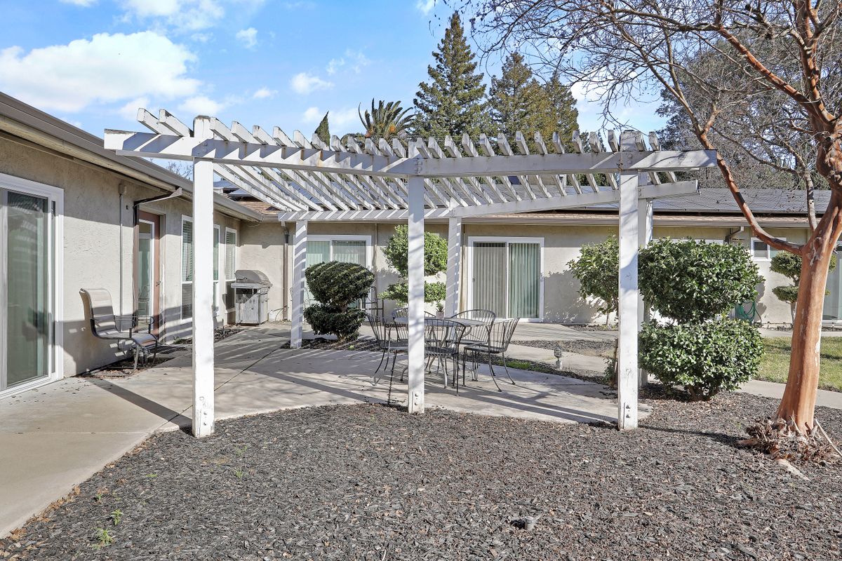 A backyard patio featuring a white pergola and outdoor seating, surrounded by landscaped greenery.