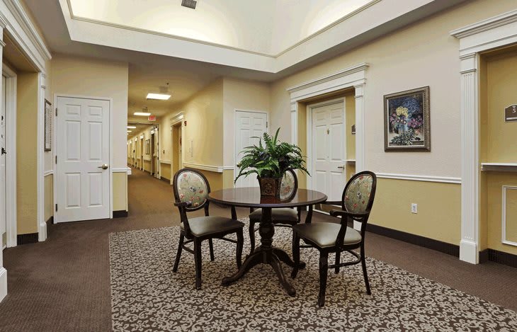 A warmly lit hallway with a table and chairs, decorated with a potted plant, surrounded by carpeted flooring and artwork on the wall.