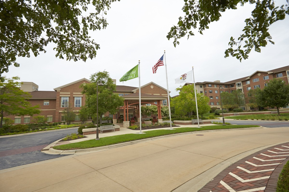 A building with three flags in front, surrounded by trees and a driveway on a cloudy day.