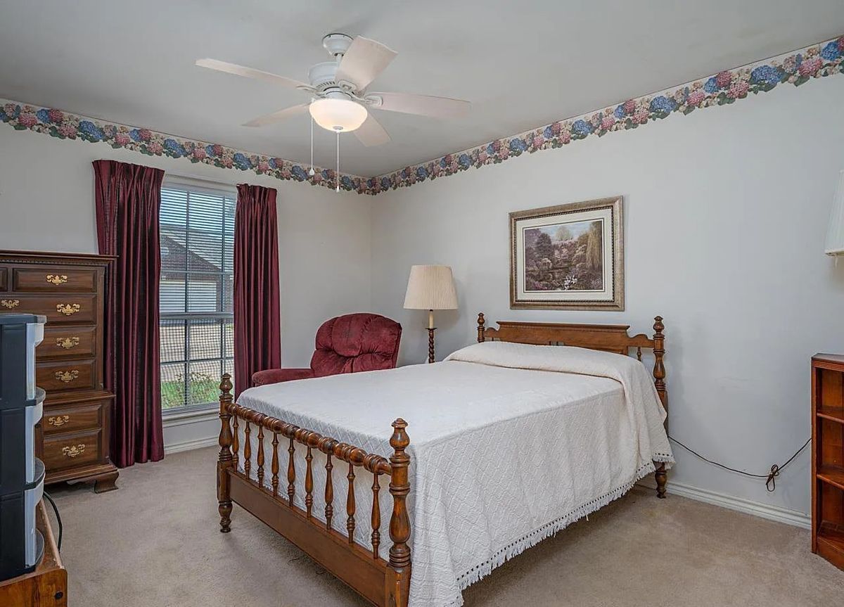 A cozy bedroom with a wooden bed frame, red upholstered chair, floor lamp, and window with red curtains.