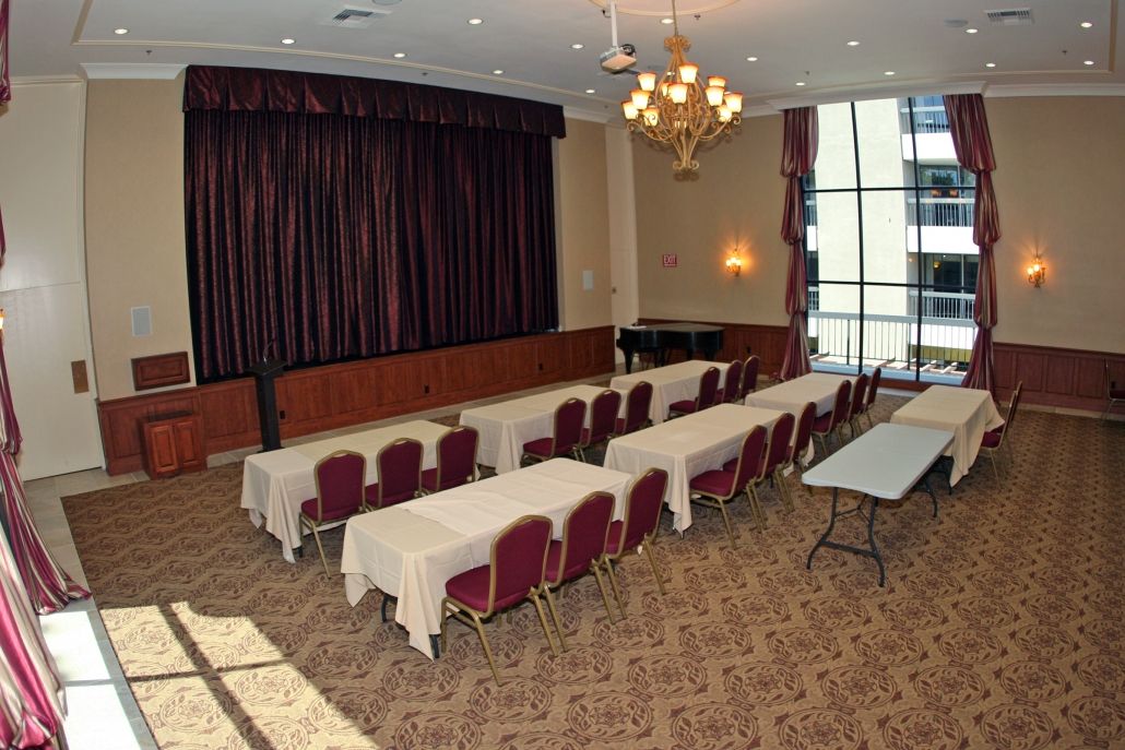 A conference room setup with rows of tables and chairs facing a stage, illuminated by a chandelier.