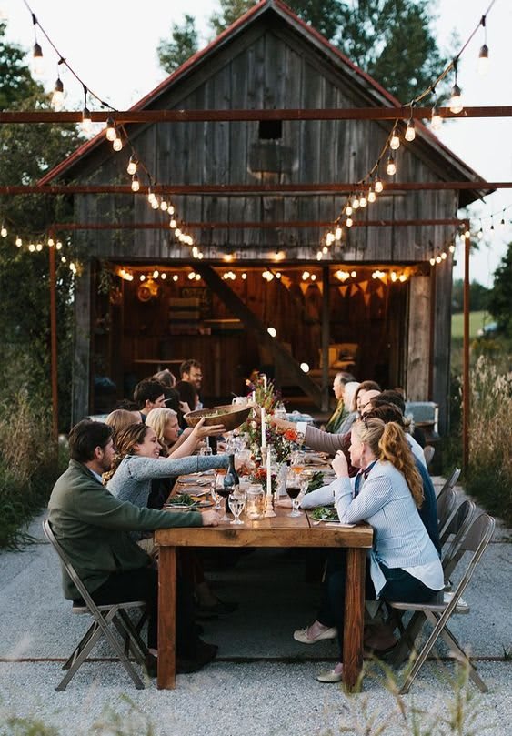 A group of people enjoying an outdoor dinner under string lights in a rustic setting.