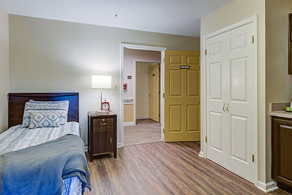 A cozy bedroom interior with a bed, nightstand, lamp, and open door leading to a hallway.