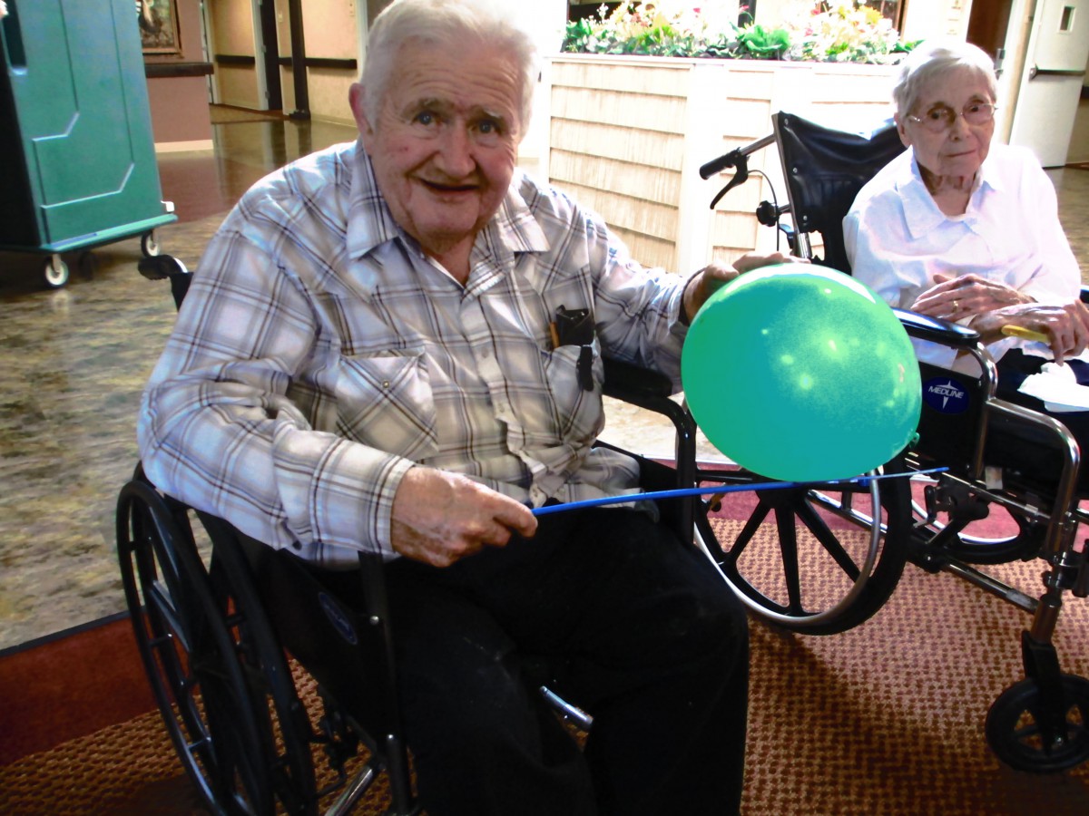 An elderly man in a wheelchair, smiling and holding a balloon indoors.
