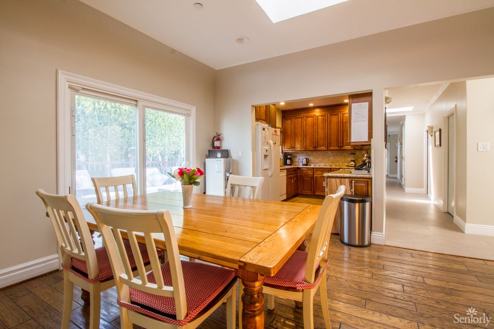 A cozy dining area with a wooden table and chairs, adjacent to a kitchen featuring warm wood cabinetry. Natural light streams in through large windows and a skylight.