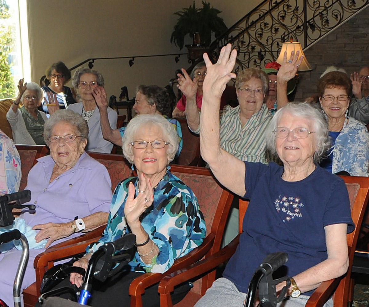 A group of elderly women sitting together indoors, smiling and waving at the camera.