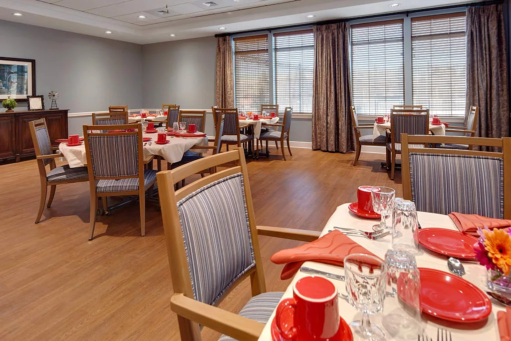 An elegantly set dining room with wooden chairs and tables, featuring red tableware and large windows.