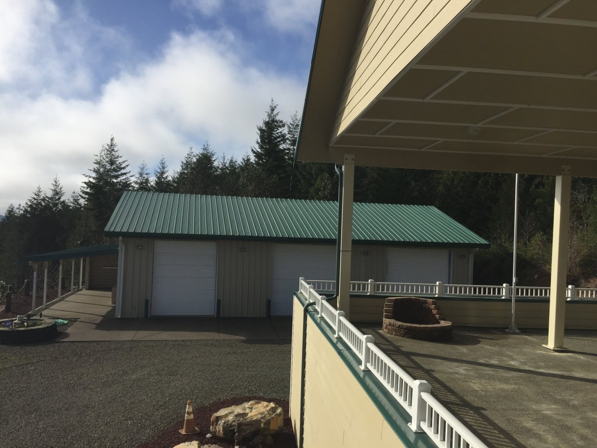 A green-roofed garage adjacent to a house with a sunny outdoor setting and trees in the background.