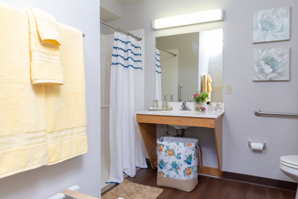 A well-lit bathroom with a sink, mirror, and shower area adorned with yellow towels and a colorful laundry basket.