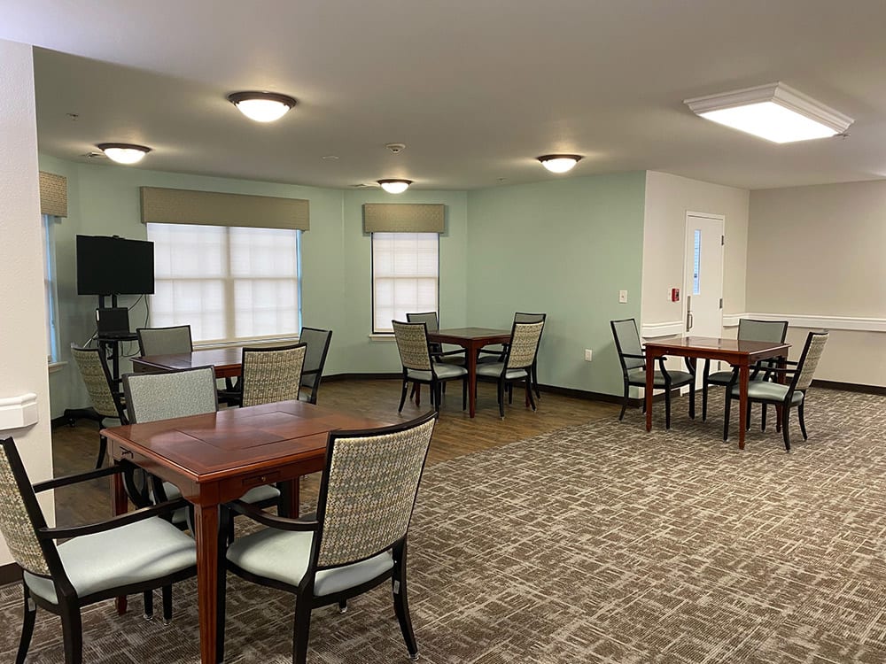 A spacious room with multiple tables and chairs, featuring carpeted flooring and windows.