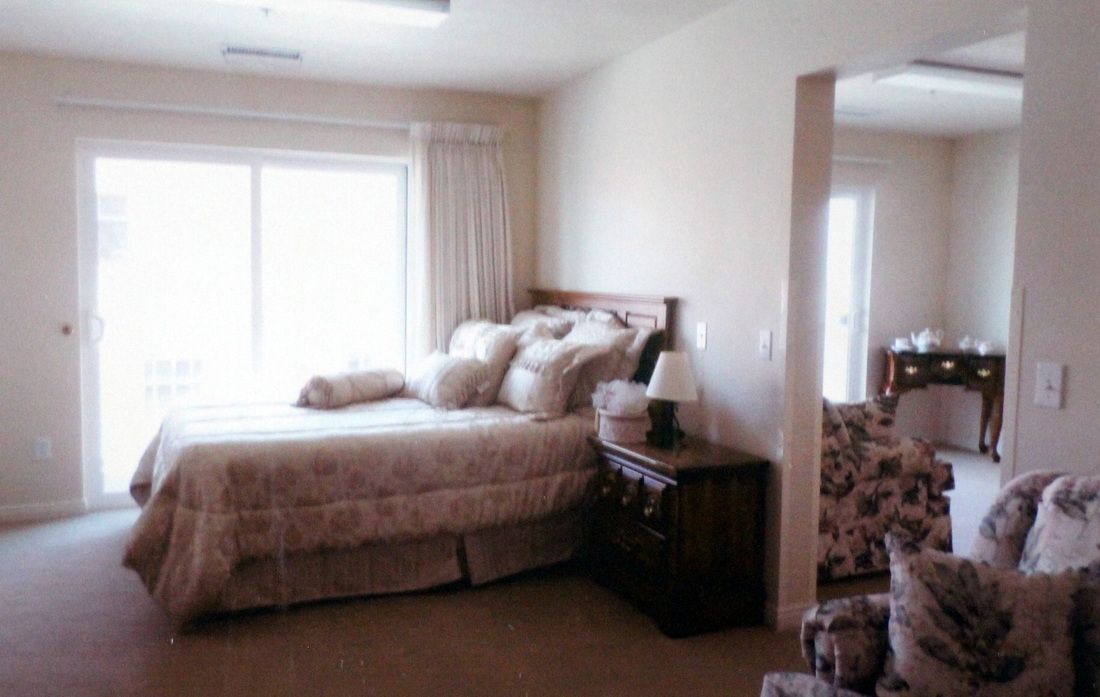 A cozy bedroom with a neatly made bed, a nightstand with a lamp, and a window letting in natural light.