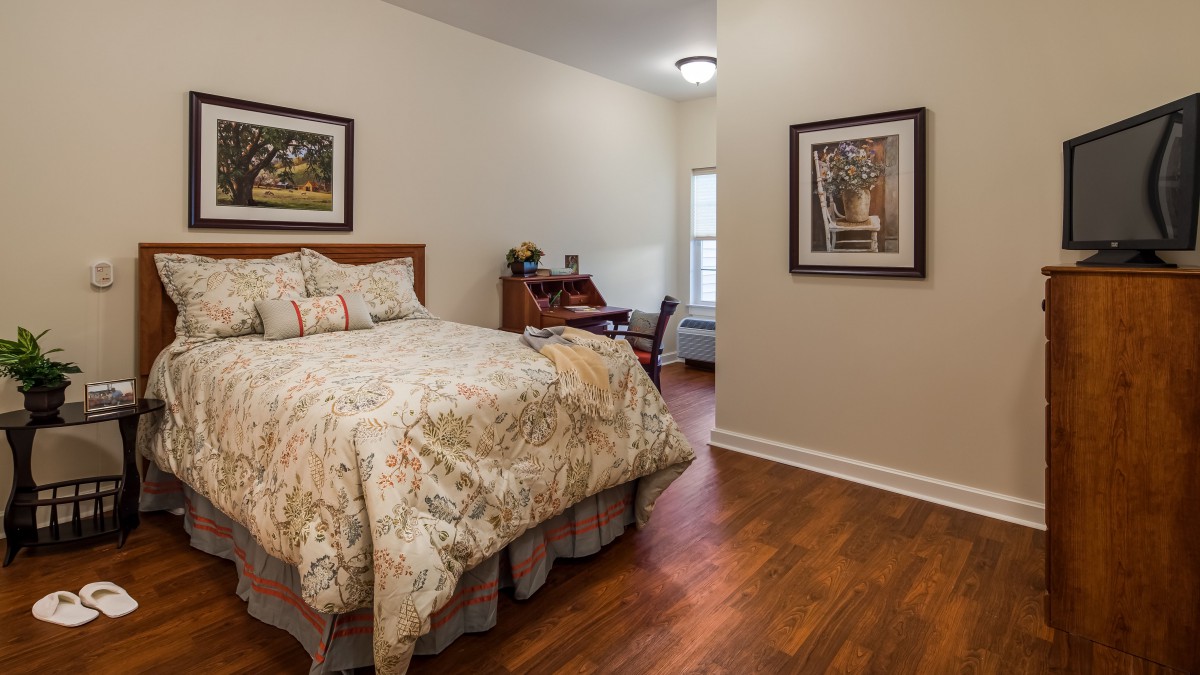 A cozy bedroom with wooden flooring, featuring a bed, artwork on the walls, and a desk near the window.
