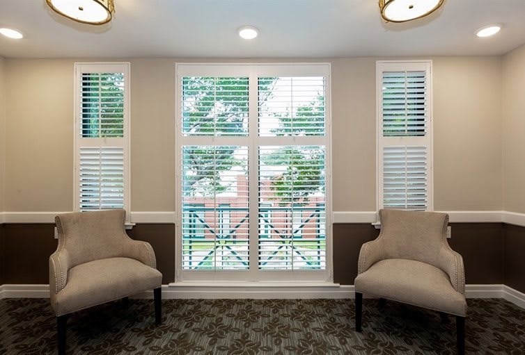 A cozy seating area with two chairs facing large windows with blinds, carpeted flooring, and ceiling lights.