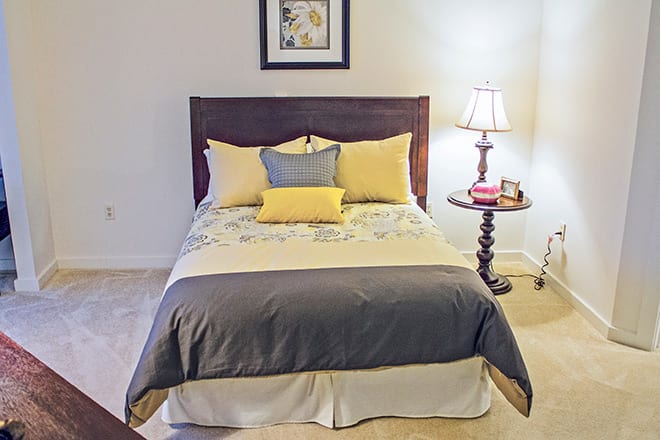 A cozy bedroom with a neatly made bed, featuring yellow and gray pillows, a decorative lamp on the nightstand, and a framed picture above the headboard.