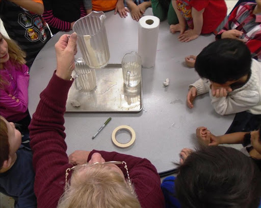 A teacher demonstrates a science experiment while children observe eagerly in a classroom setting.