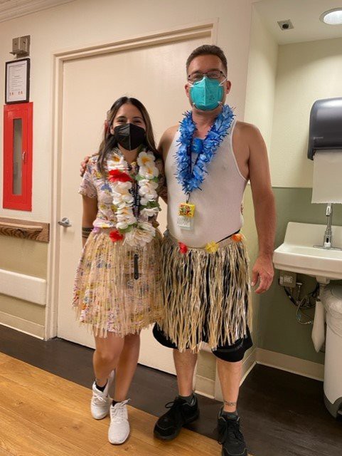 Two people in Hawaiian-themed costumes and masks pose in a hospital setting.