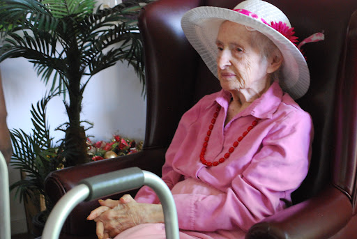 An elderly woman in a pink outfit and wide-brimmed hat sits thoughtfully in a cozy indoor setting.