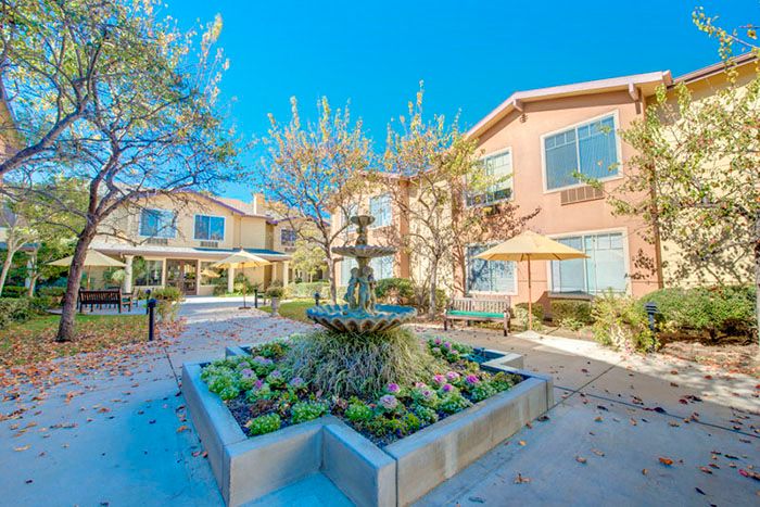 A sunny courtyard with a central fountain surrounded by flowers and trees, adjacent to residential buildings.