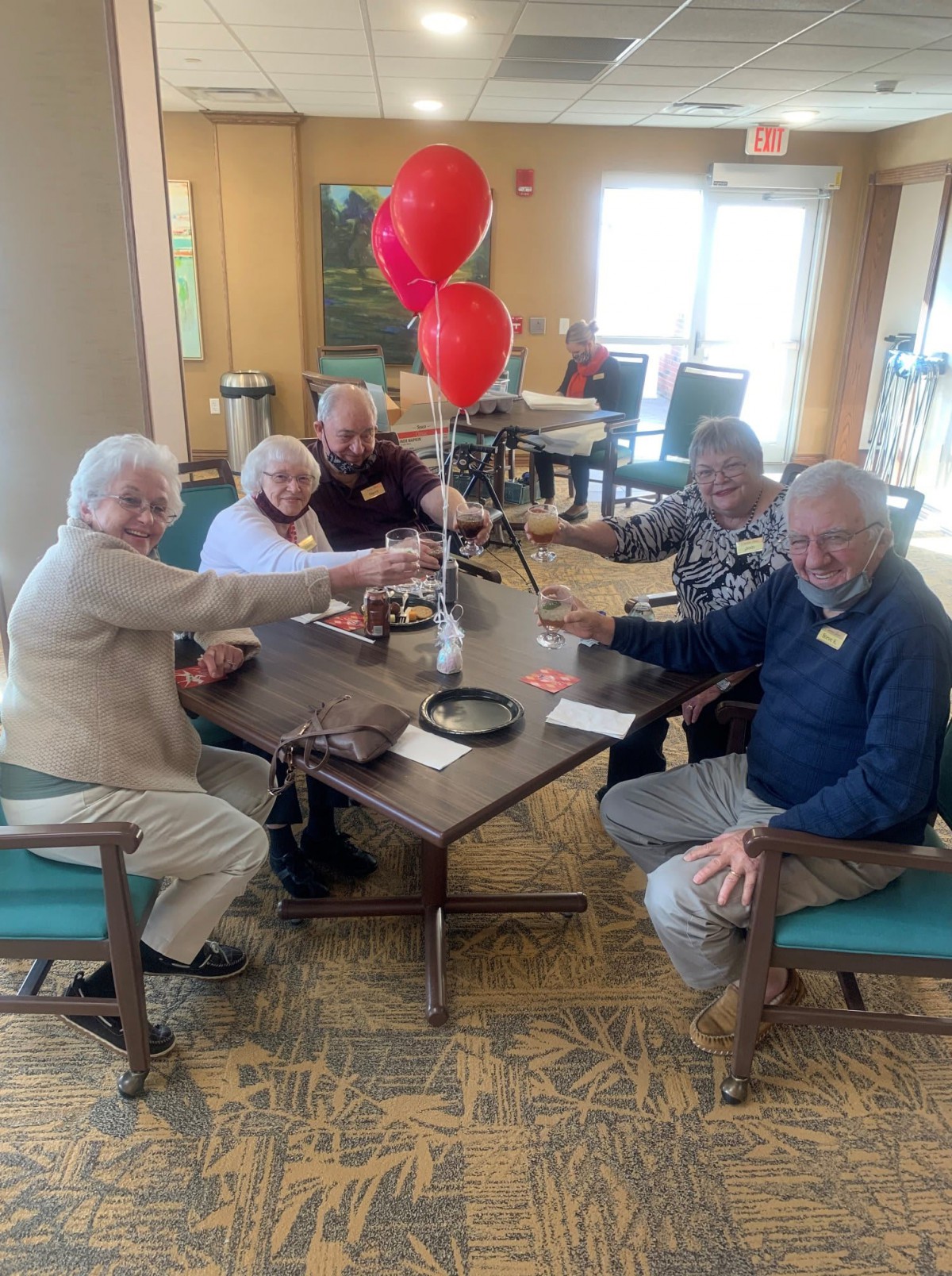 A group of seniors enjoying a celebration indoors, raising glasses with red balloons in the background.