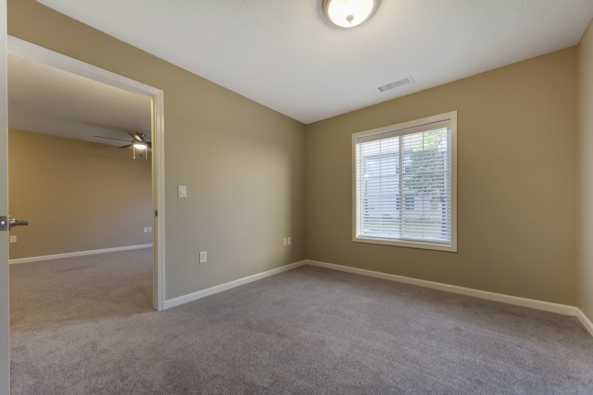 An empty room with a window, beige walls, and carpeted flooring.