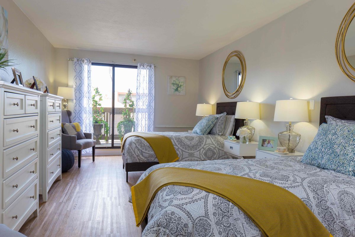 A bright and modern bedroom with two double beds, featuring yellow and gray bedding, abundant natural light, and contemporary decor.