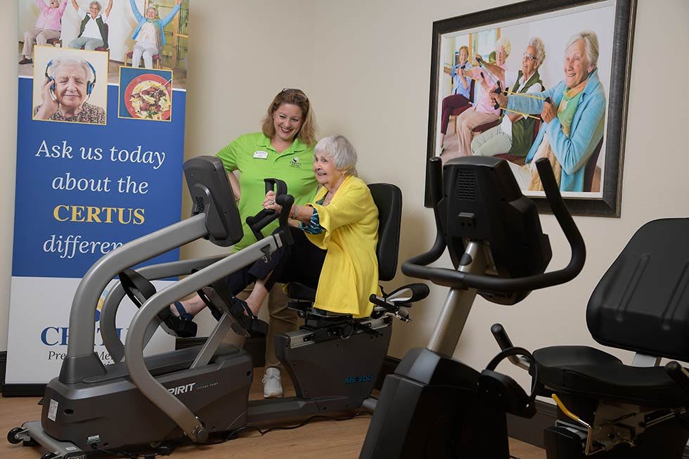A senior woman exercises on a stationary bike with assistance from a trainer in a gym setting.