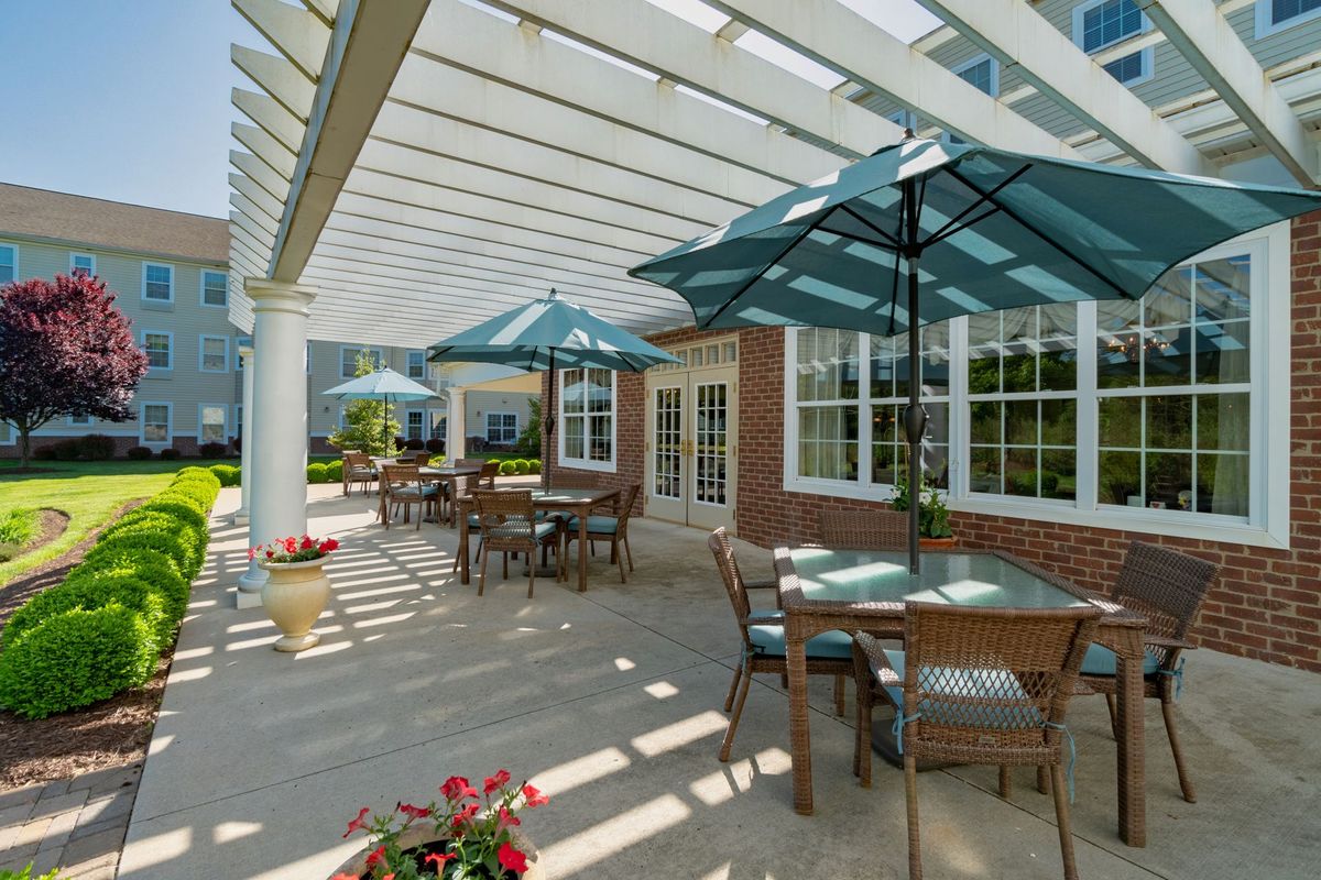 An outdoor patio area with tables, chairs, and umbrellas under a pergola beside a brick building.