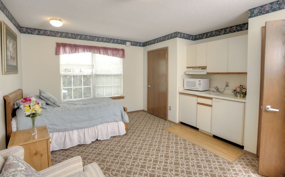 A cozy bedroom with a kitchenette, featuring a bed, microwave, and large window.