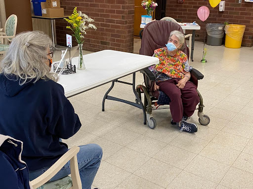 An elderly woman in a wheelchair engages in conversation with another person, both wearing masks during a social visit.