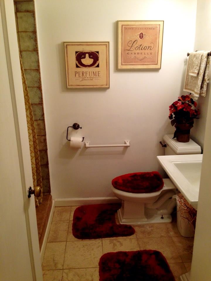 A cozy bathroom with wall art, a toilet adorned with matching red bath mats, and decorative flowers on the sink.