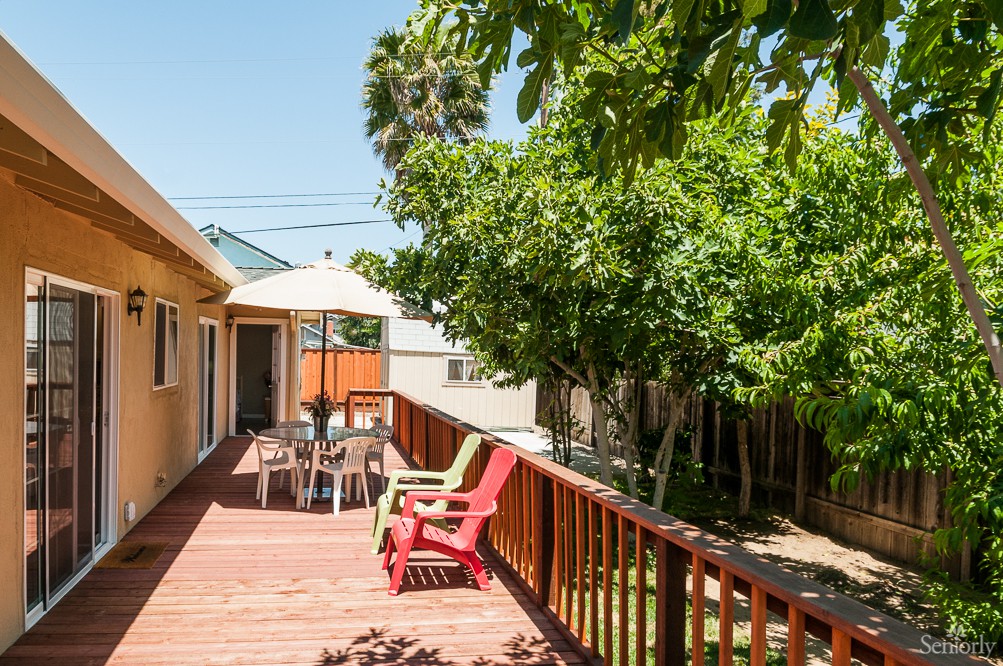 A sunny backyard deck with colorful chairs and a shaded table under lush green trees.