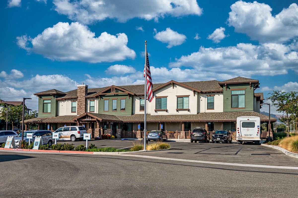 A two-story building with a parking lot, featuring parked cars and an American flag under a cloudy sky.