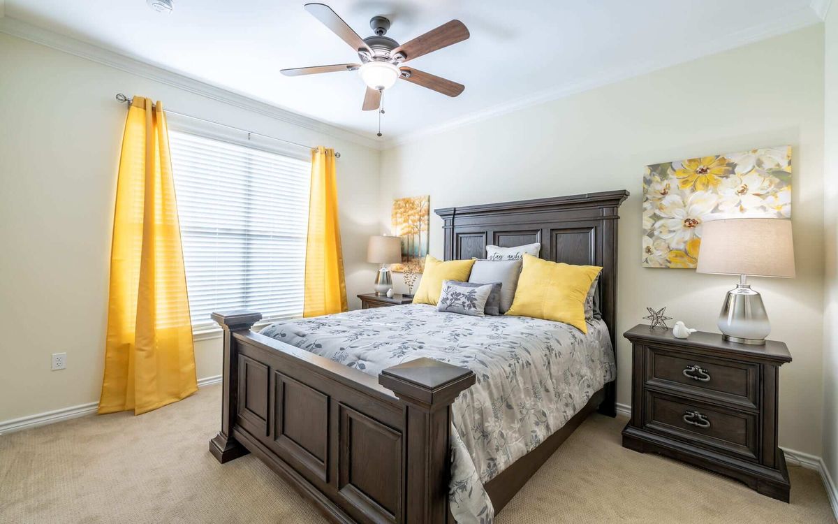 A cozy bedroom featuring a dark wood bed frame with yellow accent pillows and curtains, complemented by a ceiling fan.