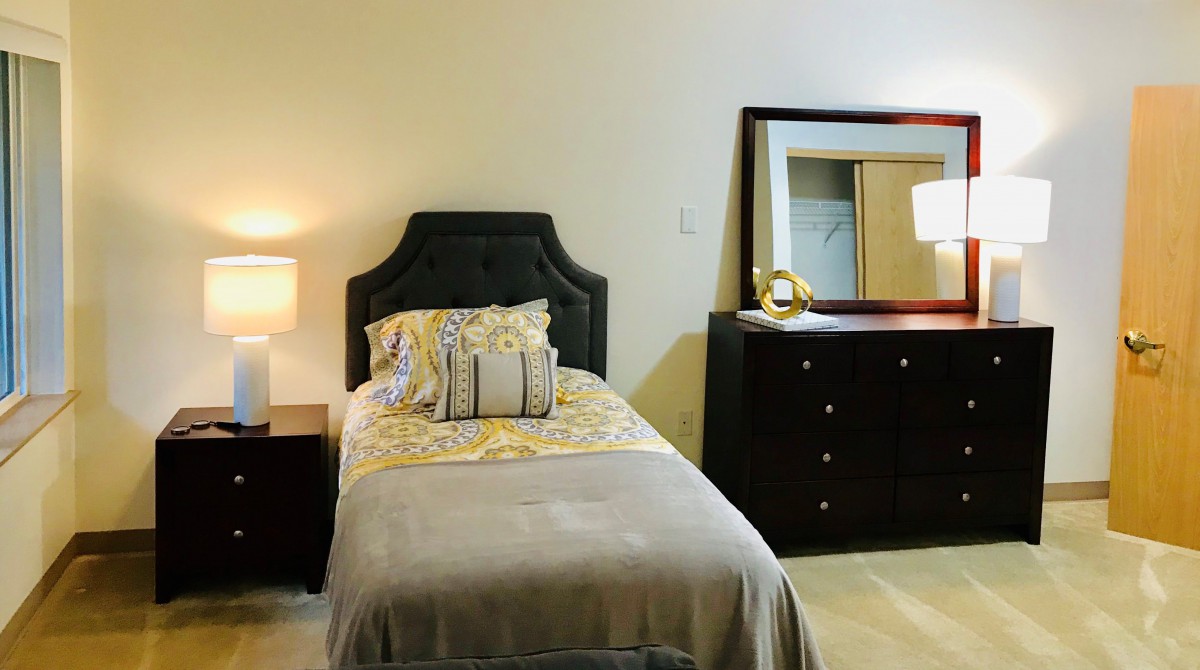 A cozy bedroom with a single bed, nightstand, lamp, and a dresser with a mirror.