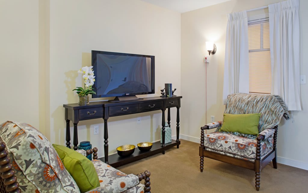 A cozy living room with a television on a console table, surrounded by floral-patterned furniture and green accent pillows.