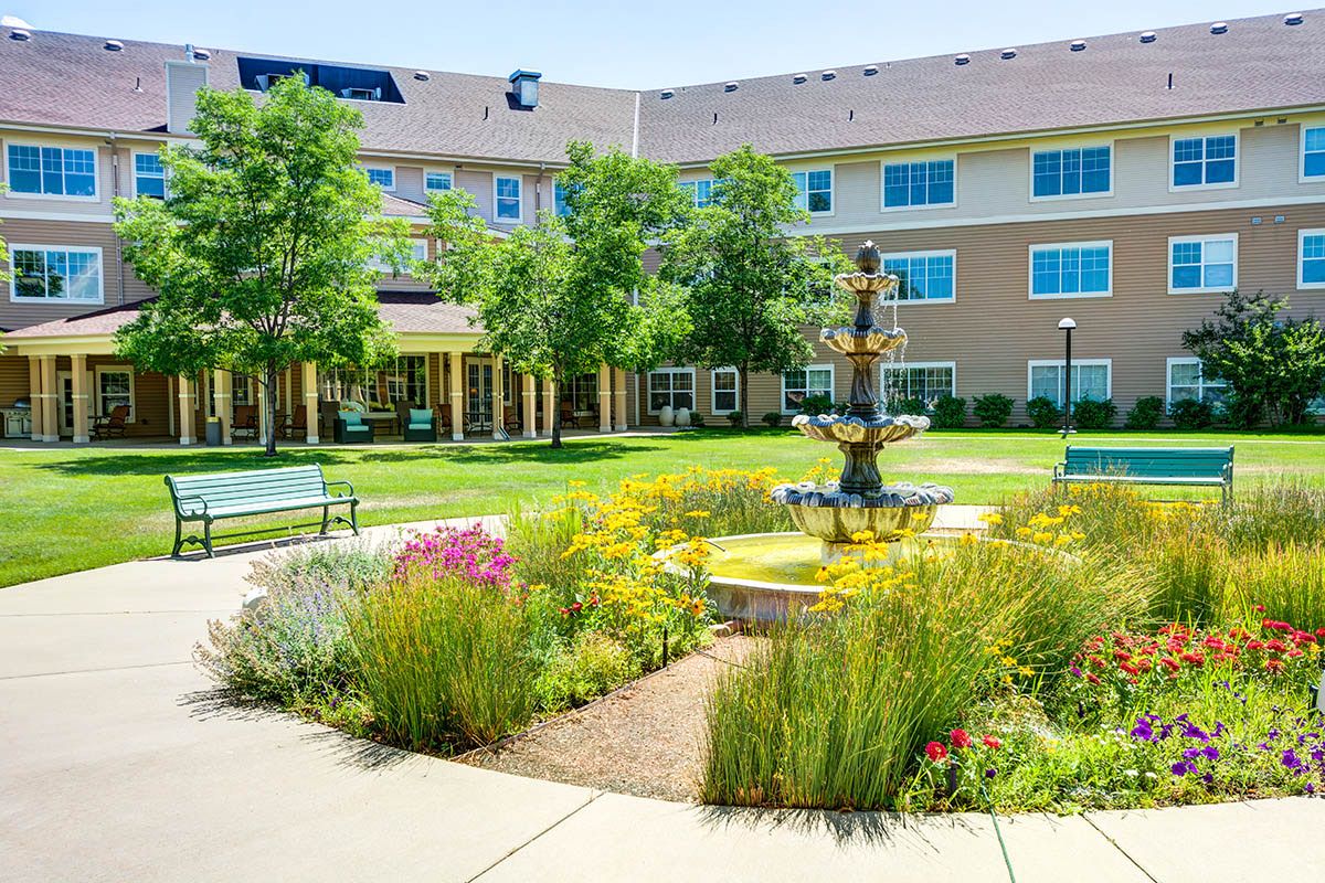 A beautifully landscaped courtyard with a central fountain, surrounded by greenery and benches in front of a multi-story building.